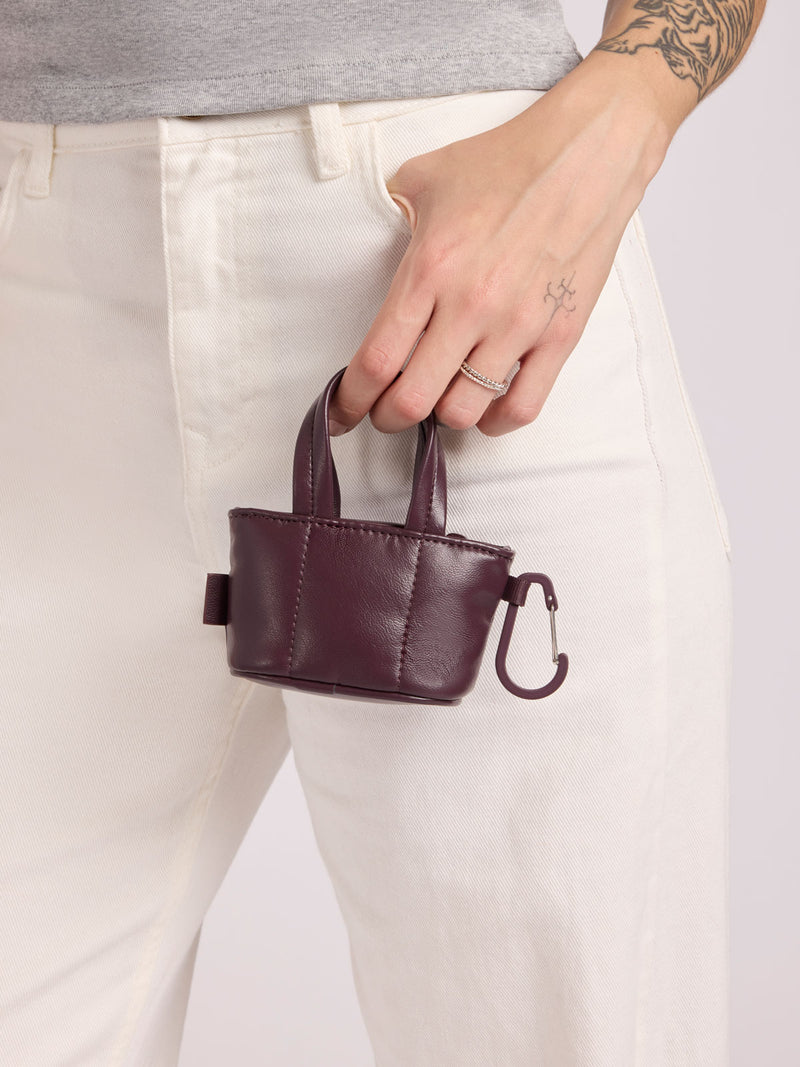 A person wearing off-white pants and a gray top holds a small Dark Cherry CALPAK Luka Tote Zip Bag Charm in Vegan Leather with top handles and a carabiner clip