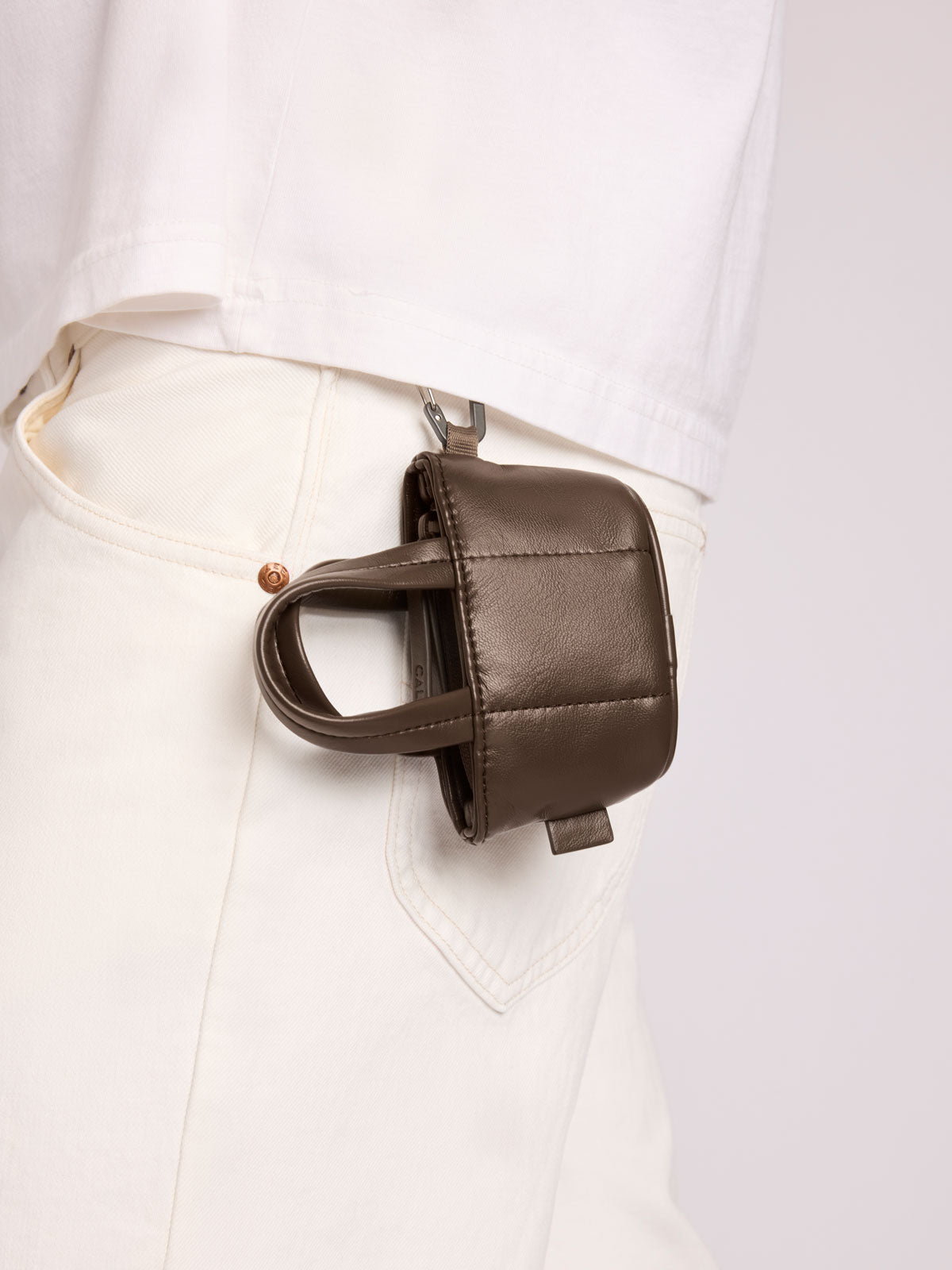 A small CALPAK Luka Tote Zip Bag Charm in Mushroom Vegan Leather with a top handle, clipped to the waist belt of a person wearing white pants and a white shirt; AMT2601-MUSHROOM-PU