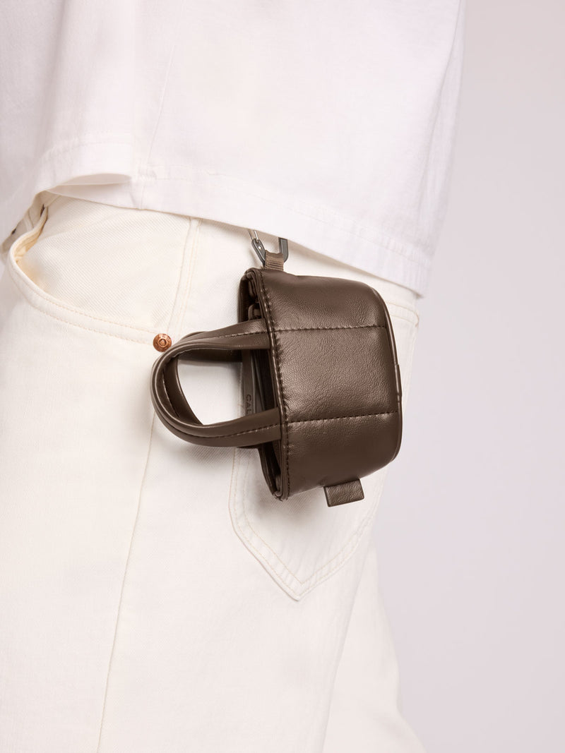 A small CALPAK Luka Tote Zip Bag Charm in Mushroom Vegan Leather with a top handle, clipped to the waist belt of a person wearing white pants and a white shirt