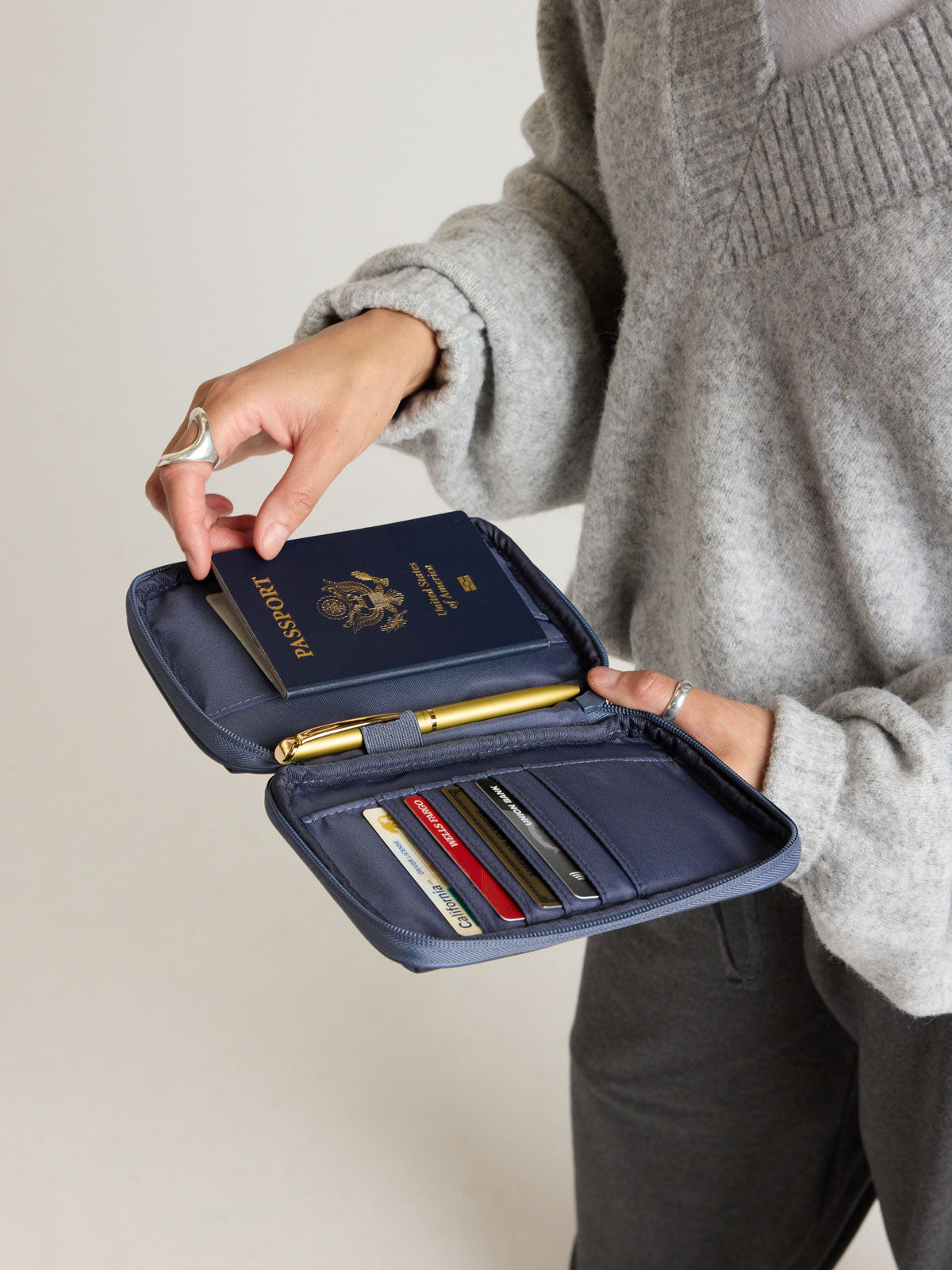 Model putting passport into the CALPAK Luka Zippered Passport Wallet in dark blue metallic material in Metallic Indigo; PZW2301-METALLIC-INDIGO