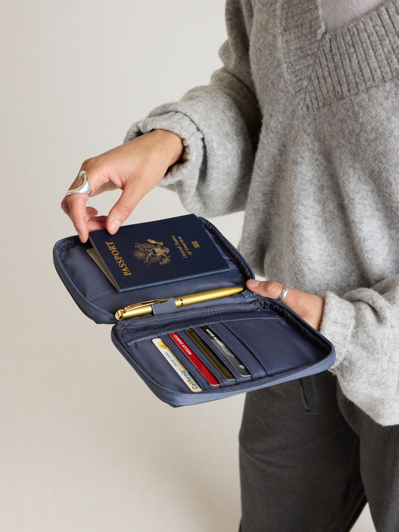 Model putting passport into the CALPAK Luka Zippered Passport Wallet in dark blue metallic material in Metallic Indigo