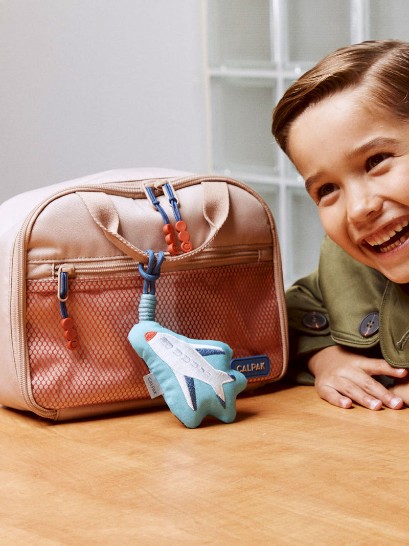 CALPAK Kids Lunch Bag in brown and tan Khaki with Airplane Pluffy Bag Charm with child smiling next to it