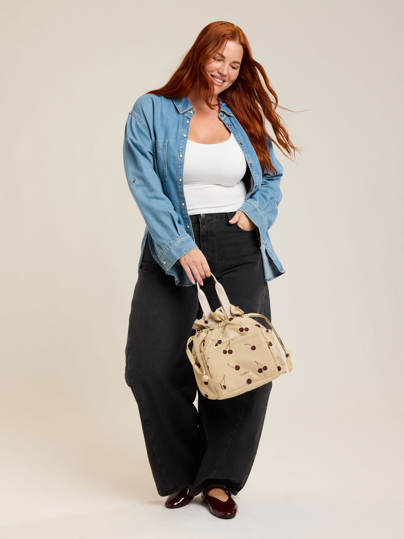 Model with CALPAK Insulated Lunch Bag in Oatmeal Cherry in tan with cherry print with drawstring opened
