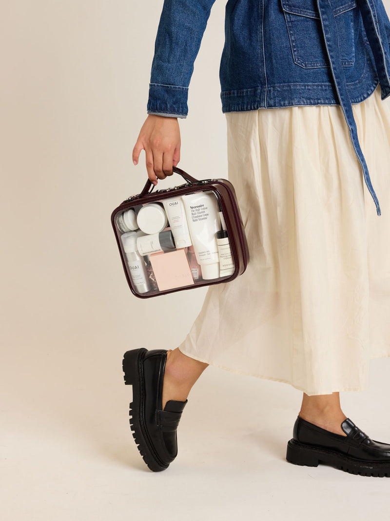 Model carrying CALPAK Medium Clear Cosmetic Case in maroon Glazed Dark Cherry on a white background