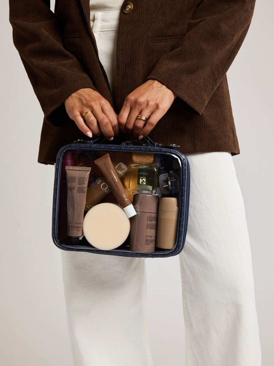 A person wearing a brown corduroy blazer and white pants holds a Medium Clear Cosmetics Case in Navy croc-embossed texture against a light gray studio background; CMM2201-NAVY-FAUX-CROC