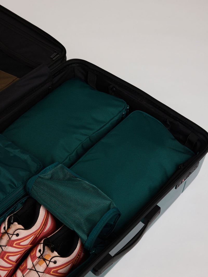 Two dark green teal Kale CALPAK Medium Compression Packing Cubes arranged inside an open black suitcase, with a pair of pink and black sneakers visible in the lower left compartment