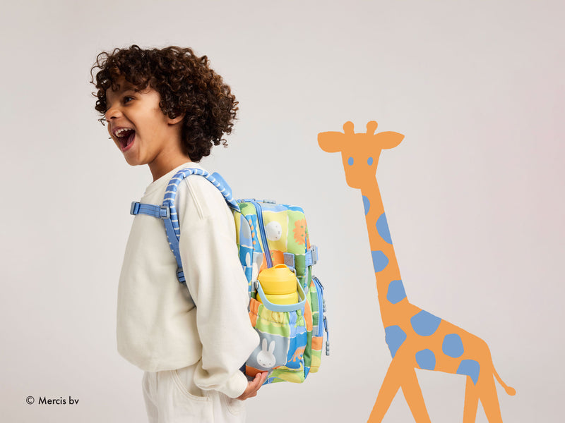 A child with curly brown hair wearing a white sweatshirt and white pants, smiling and looking back over their shoulder while wearing a colorful patterned school backpack. To the right is a large orange and blue giraffe wall decal on a light gray background.