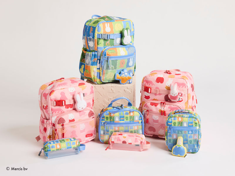 A collection of CALPAK + Miffy pastel-colored school bags and small pouches with bunny motifs, arranged on a light beige surface against a white background. The set includes pink and blue bags in various sizes, some stacked on top of each other.