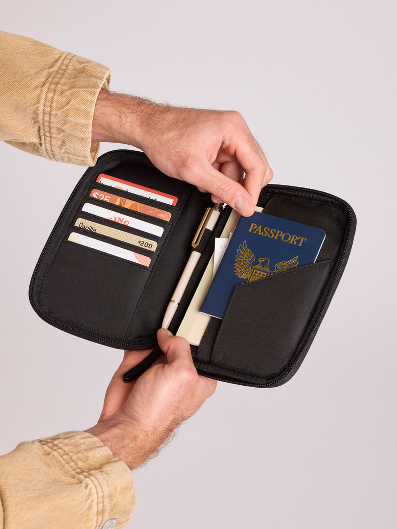 A CALPAK Luka Passport Wallet in Black Vegan Leather shown open, held by a person's hands. Inside, card slots hold credit cards, a pen, and a blue passport is tucked into the side pocket