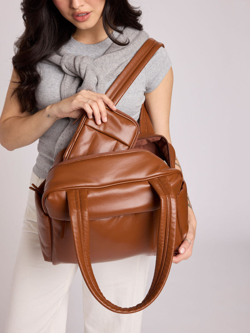 A woman wearing a gray knit top and white pants holds a CALPAK Luka ZIp Passport Wallet in Honey Brown Vegan Leather with matching Luka Mini Duffel against a light gray studio background