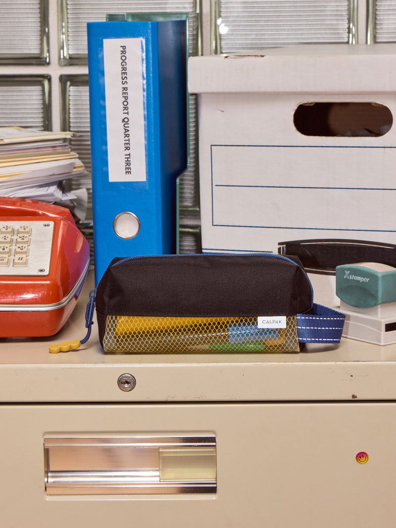 CALPAK Kids Pencil Case in Black front view with blue zippers and yellow mesh front pocket on a school cabinet
