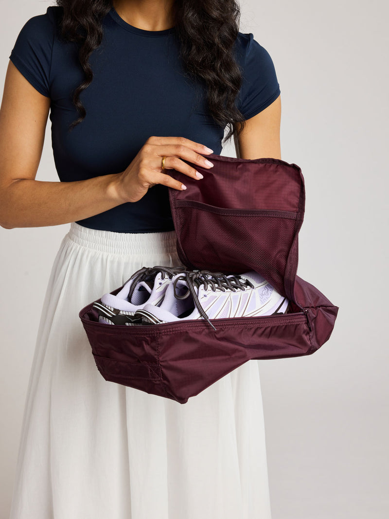 A woman wearing a navy blue top and white skirt holds an burgundy Dark Cherry rectangular CALPAK shoe cube with zippers and top handle shown on a plain background