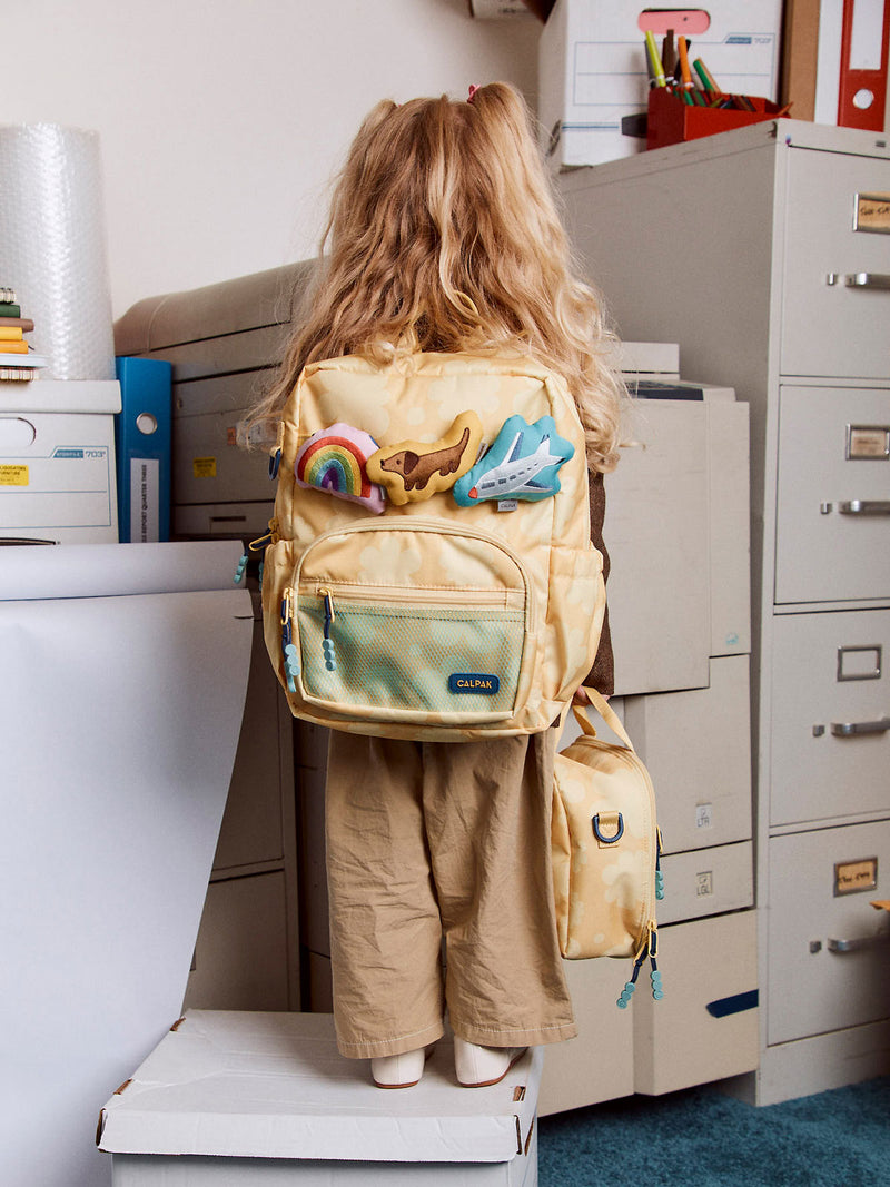 CALPAK Kids Small Backpack in yellow floral print Yellow Bloom being worn by student with Pluffy Bag Charms