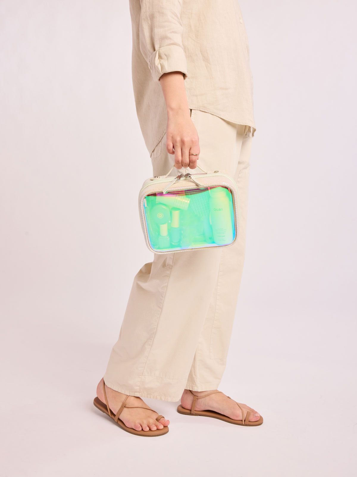 Person wearing beige holding a CALPAK Small Clear Cosmetic Case in Moonbeam with white border and transparent holographic iridescent makeup bag with pink on a white background; CCM2001-MOONBEAM