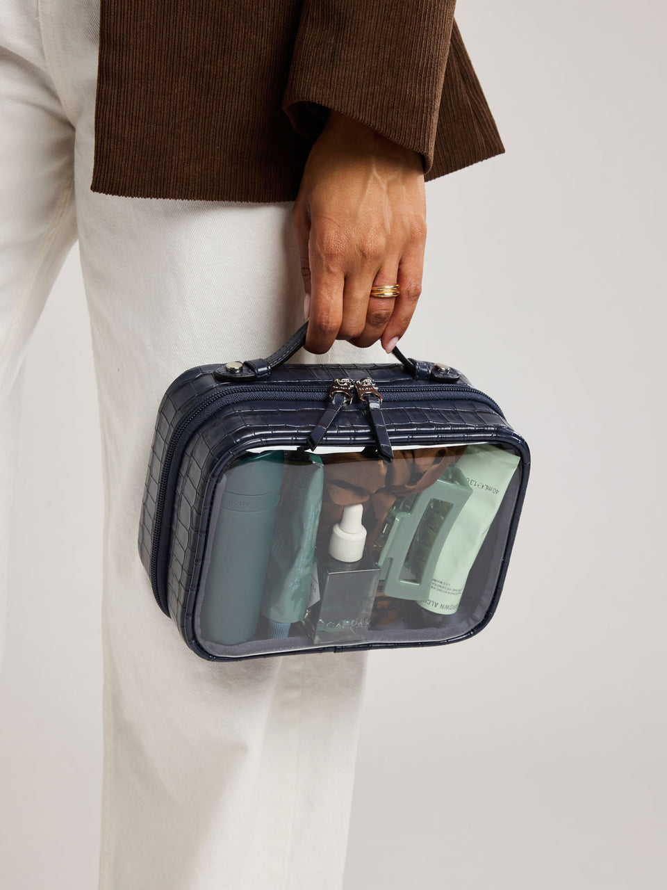 A person in a brown corduroy jacket and white pants holds a navy blue Small Clear Cosmetics Case with a Navy blue croc-embossed finish; CCM2001-NAVY-FAUX-CROC