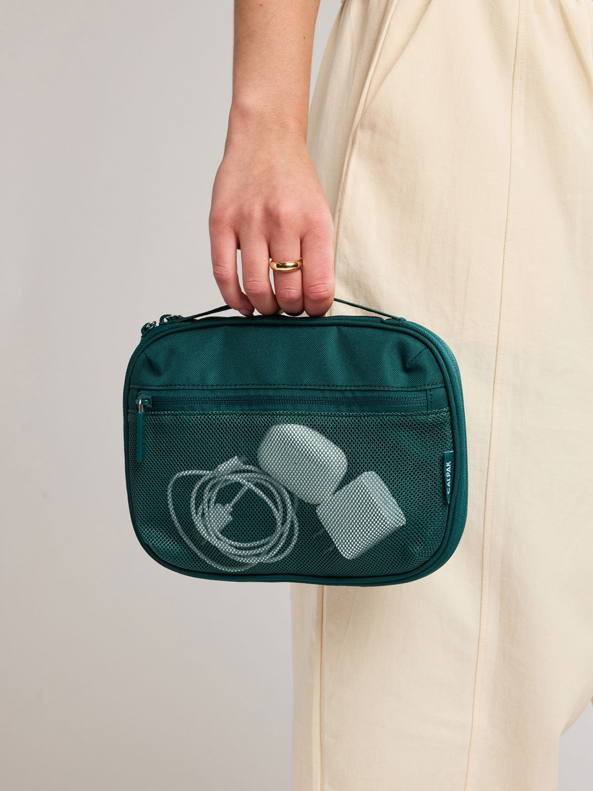 A person in a cream dress holds a dark teal green CALPAK Tech Organizer containing white earbuds and a charging cable. The person is wearing a gold ring on their finger; ATC2001-KALE