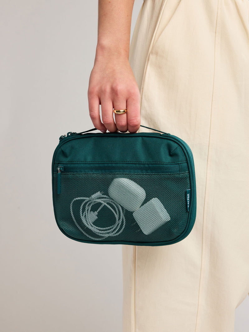 A person in a cream dress holds a dark teal green CALPAK Tech Organizer containing white earbuds and a charging cable. The person is wearing a gold ring on their finger