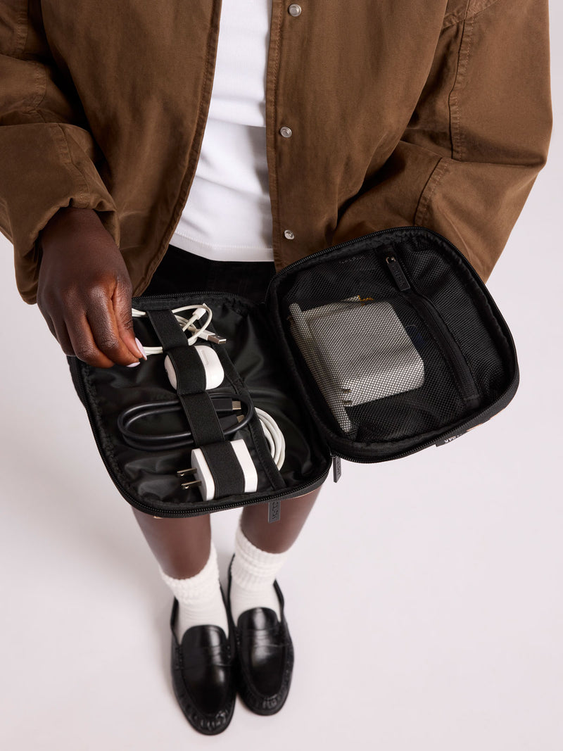 A person wearing a brown jacket and white shirt holds open CALPAK Tech Organizer pouch in Leopard containing a power strip, cables, and a gray power bank, against a light gray studio background