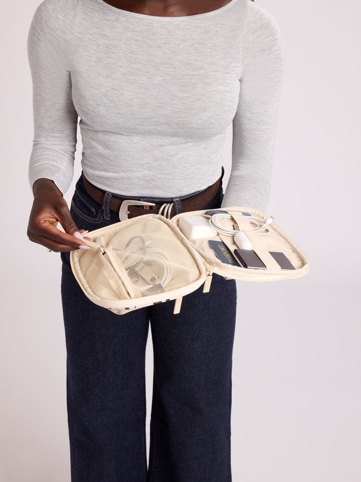 A person wearing a light gray long-sleeve top and dark blue jeans holds open a rectangular CALPAK Tech Organizer pouch in Snow Leopard with a black and beige animal print Leopard pattern, featuring a cream-colored zipper and a small black brand tag on the right side; ATC2001-SNOW-LEOPARD