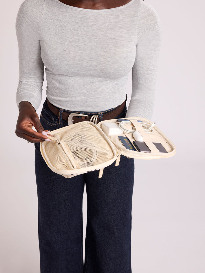 A person wearing a light gray long-sleeve top and dark blue jeans holds open a rectangular CALPAK Tech Organizer pouch in Snow Leopard with a black and beige animal print Leopard pattern, featuring a cream-colored zipper and a small black brand tag on the right side