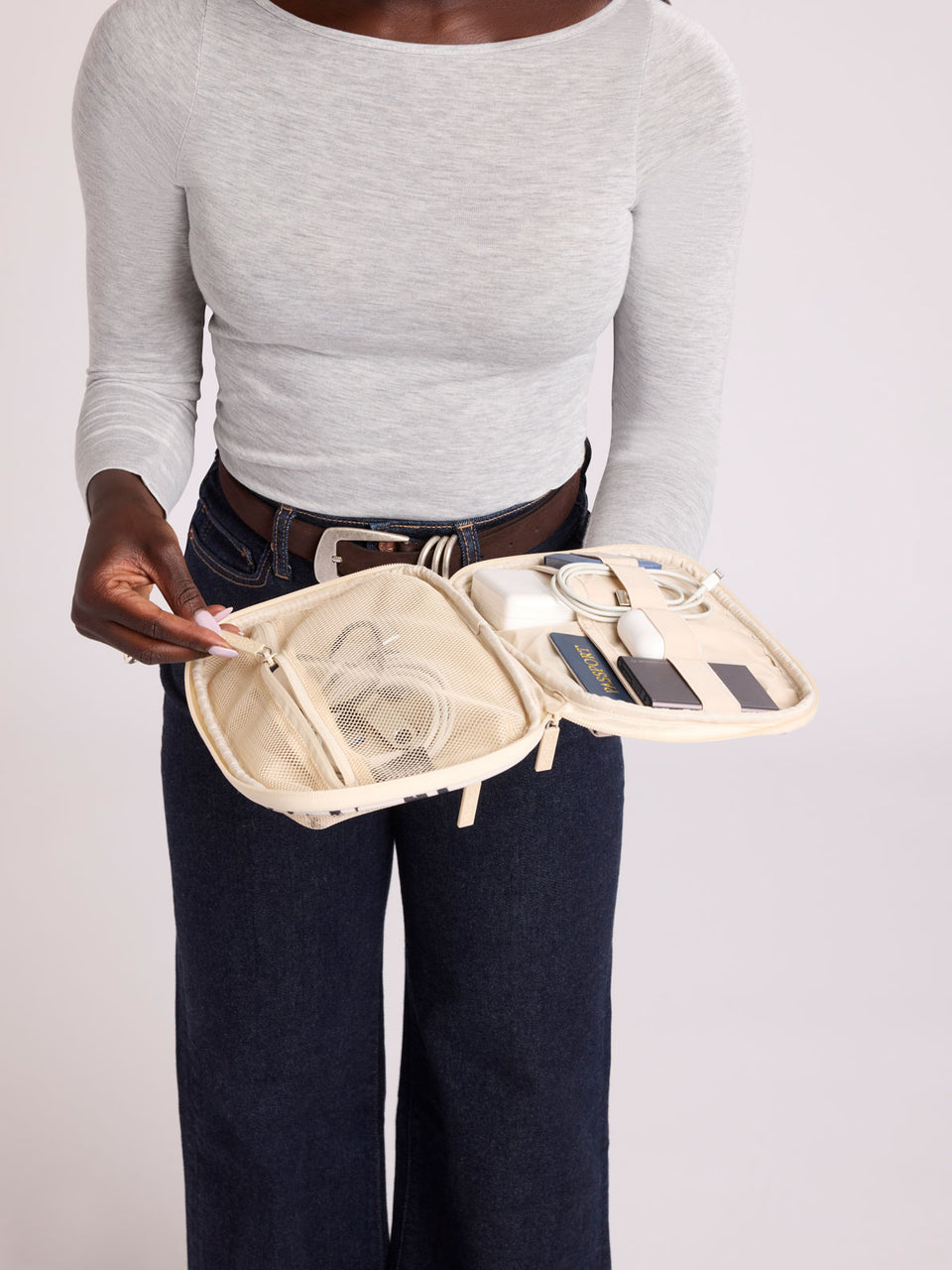 A person wearing a light gray long-sleeve top and dark blue jeans holds open a rectangular CALPAK Tech Organizer pouch in Snow Leopard with a black and beige animal print Leopard pattern, featuring a cream-colored zipper and a small black brand tag on the right side; ATC2001-SNOW-LEOPARD