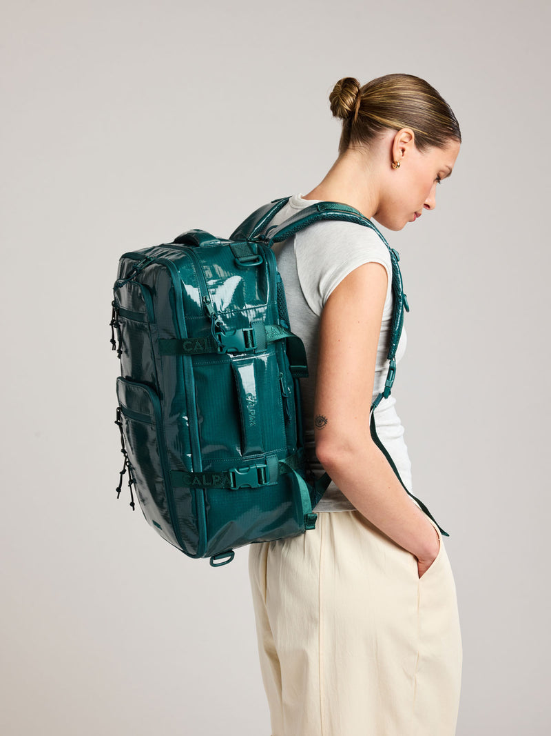 Model in a white shirt wearing the CALPAK Terra 26L Laptop Backpack in dark blue green Kale with multiple pockets on a white background