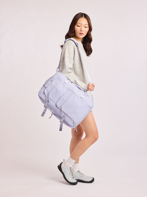 Model wearing duffel strap of CALPAK Terra 26L Laptop Duffel Backpack with multiple pockets in light purple Sea Glass on a white background; BPH2201-SEA-GLASS