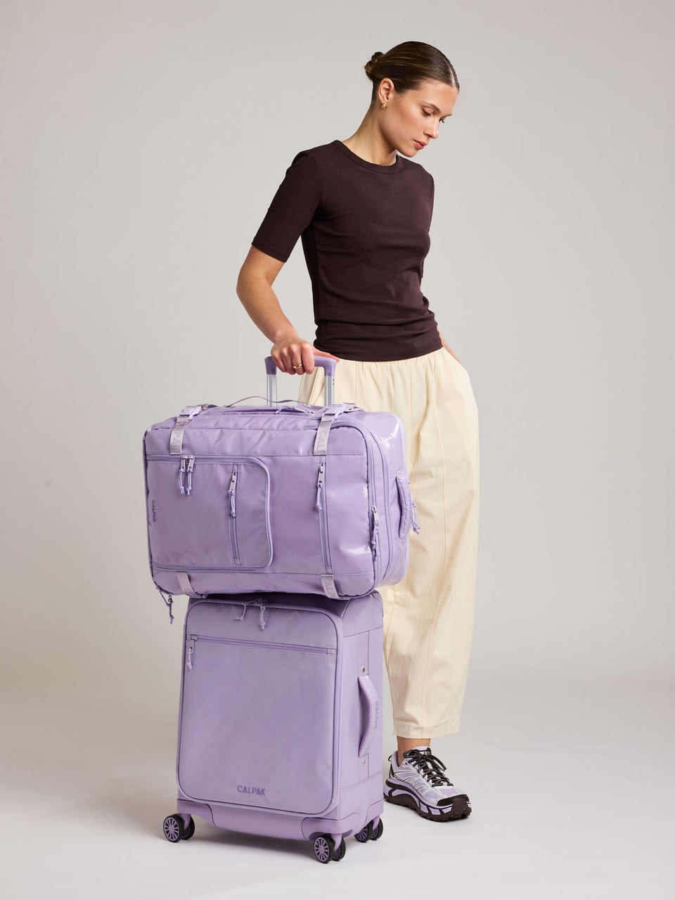 Woman with suitcase and the CALPAK Terra 35L Laptop Backpack Duffel in lavender Amethyst on top of suitcase with a white background; BPTL2601-AMETHYST