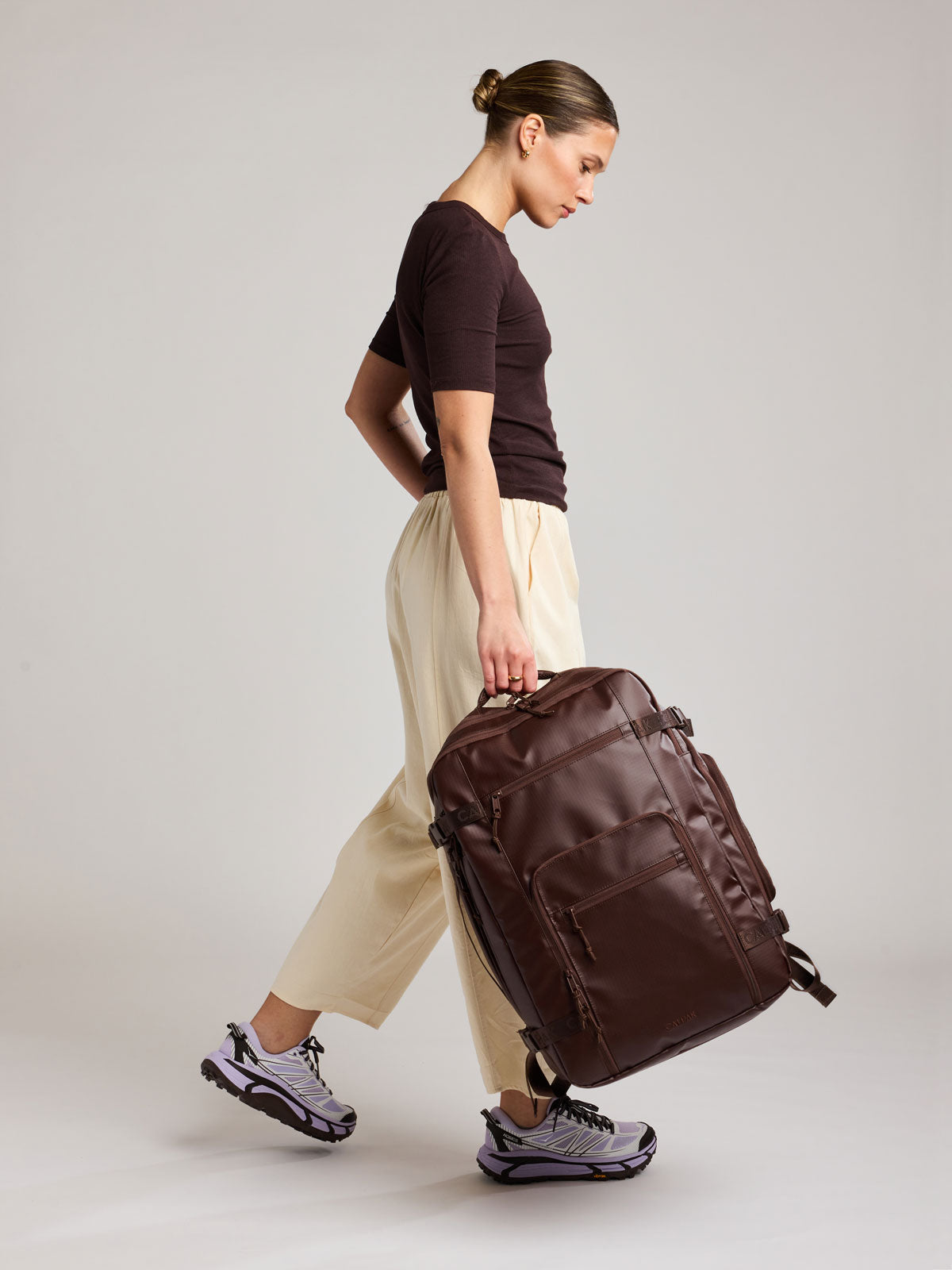 Woman carrying by the top handle the CALPAK Terra 35L Laptop Backpack Duffel in matte brown Walnut on a white background; BPTL2601-WALNUT
