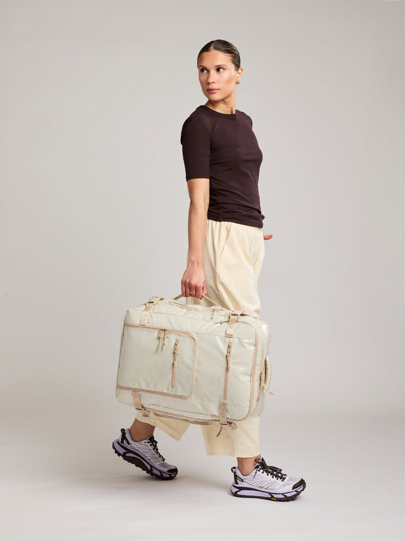 Woman holding the side handle of the CALPAK Terra 35L Laptop Backpack Duffel in off-white White Sands with tan accents on a white background