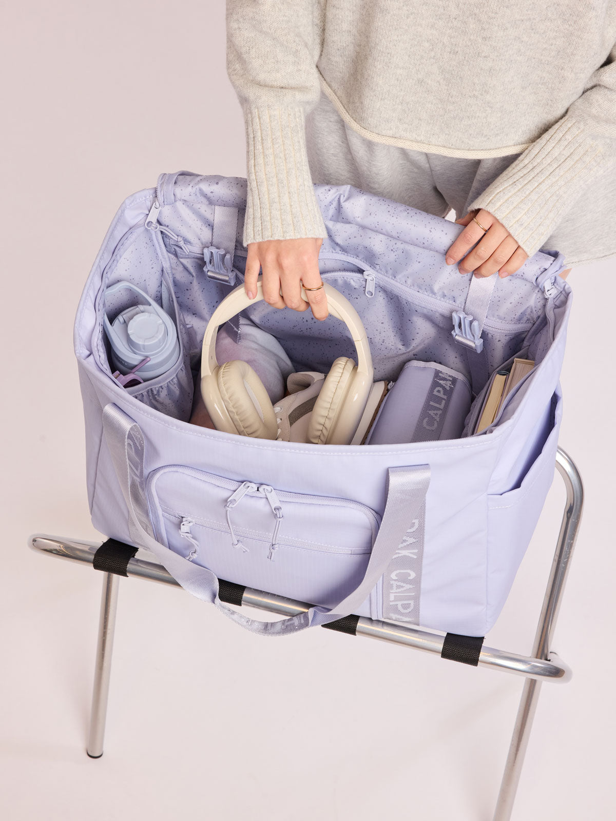Model adding headphones into the CALPAK Terra 35L Tote Bag with multiple pockets and shoulder straps in shiny light lavender Sea Glass on a white background; TTO2401-SEA-GLASS