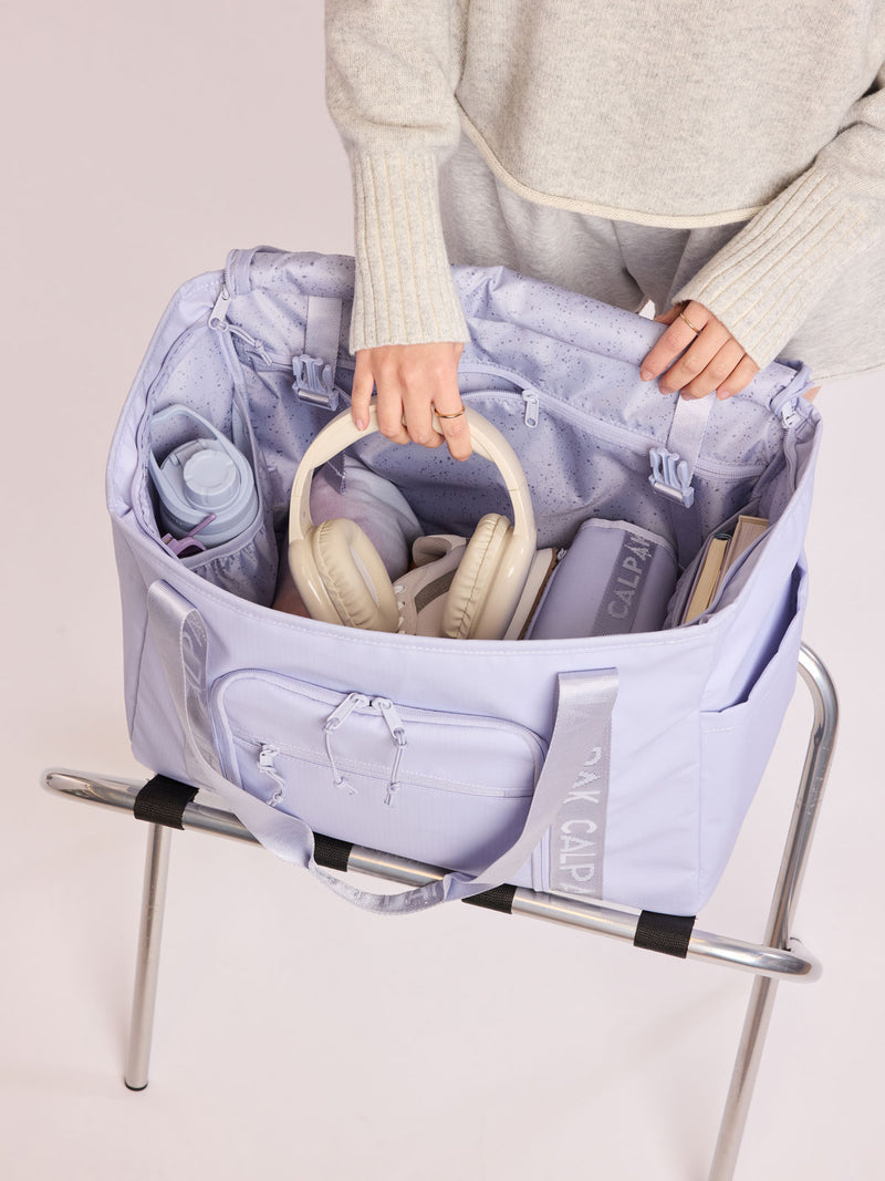 Model adding headphones into the CALPAK Terra 35L Tote Bag with multiple pockets and shoulder straps in shiny light lavender Sea Glass on a white background