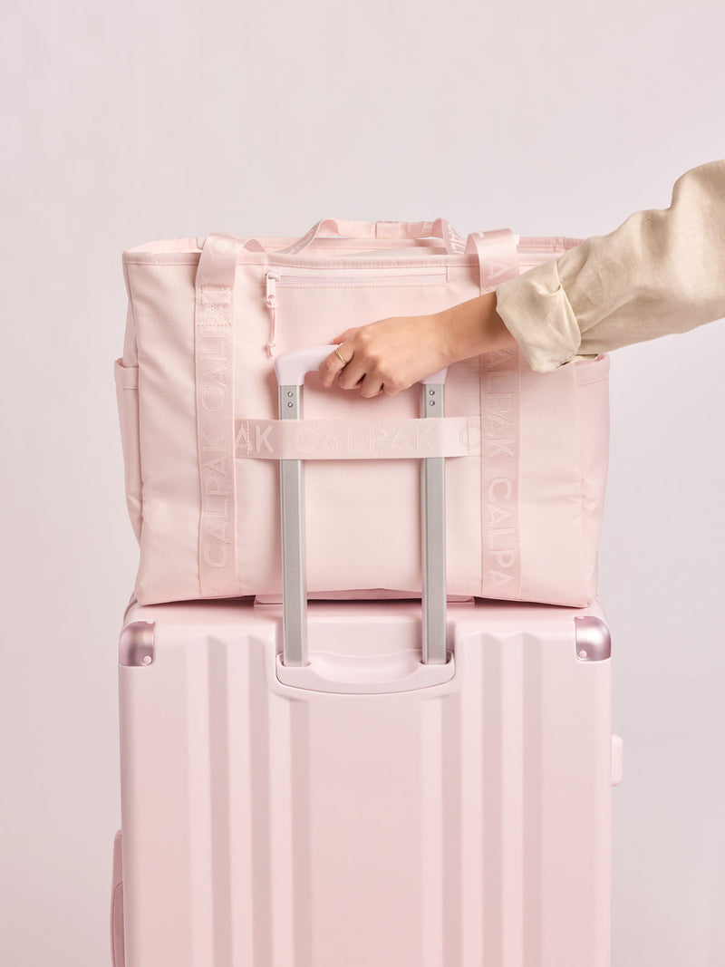 Model holding a luggage with the CALPAK Terra 35L Tote Bag with multiple pockets and shoulder straps in shiny light pink Shell Pink on a white background and luggage trolley sleeve