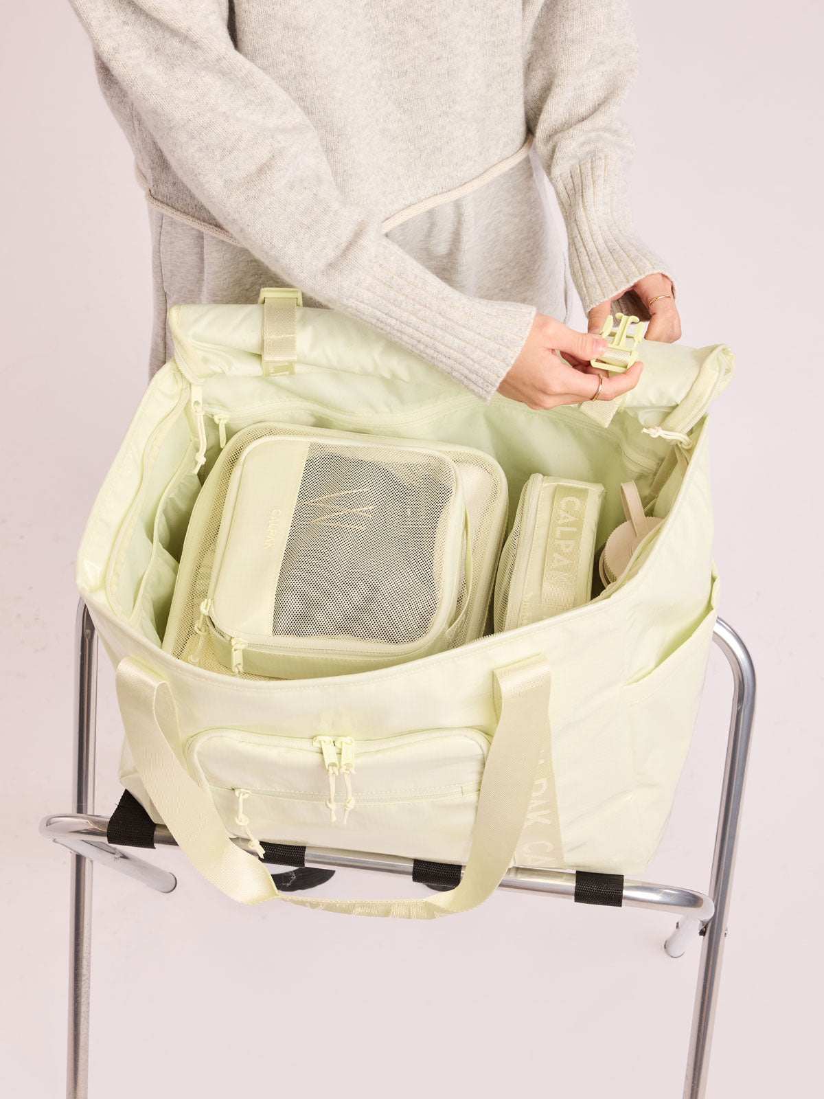 Model adding the small and medium packing cubes of CALPAK Terra 5-Piece Packing Cube Set in light green Celadon with two small cubes, two medium cubes, and one large mesh cube with zippers and mesh into a bag; PCT2601-CELADON