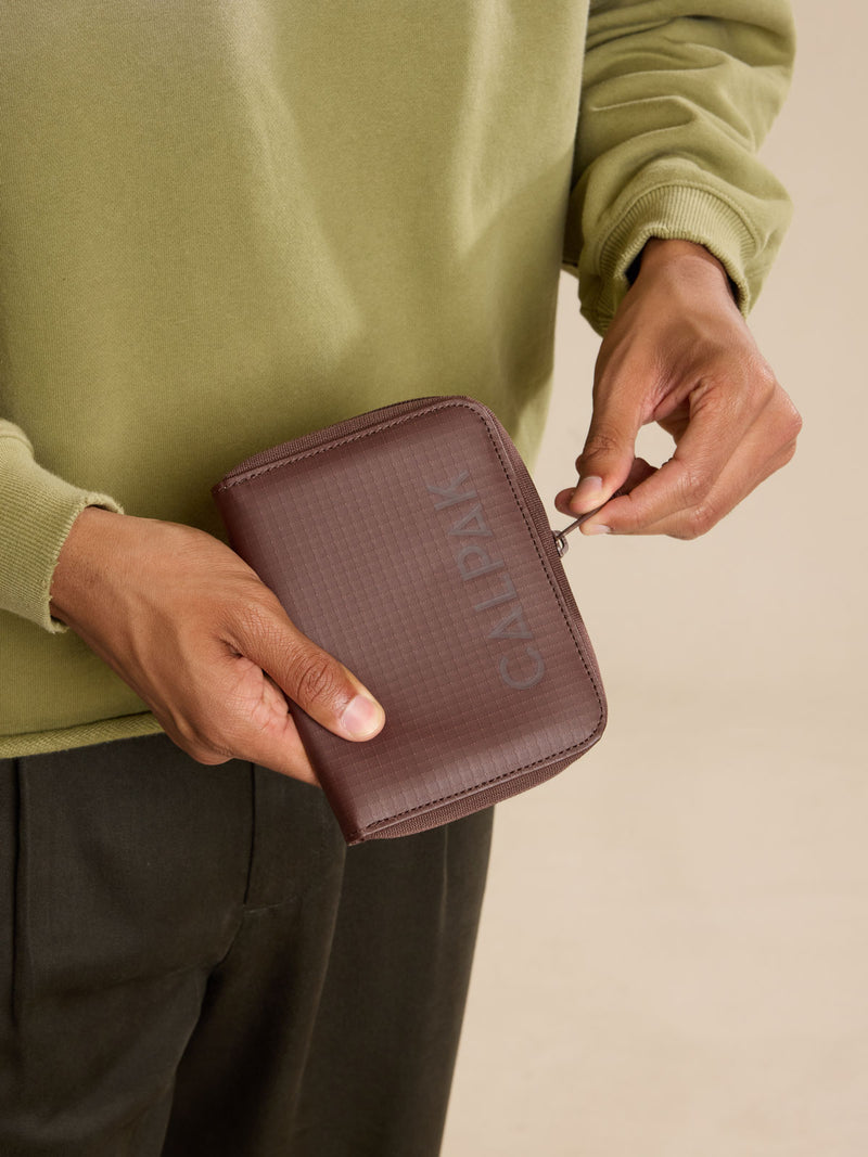 Model zipping up the CALPAK Terra Zippered Passport Wallet in brown Walnut front view