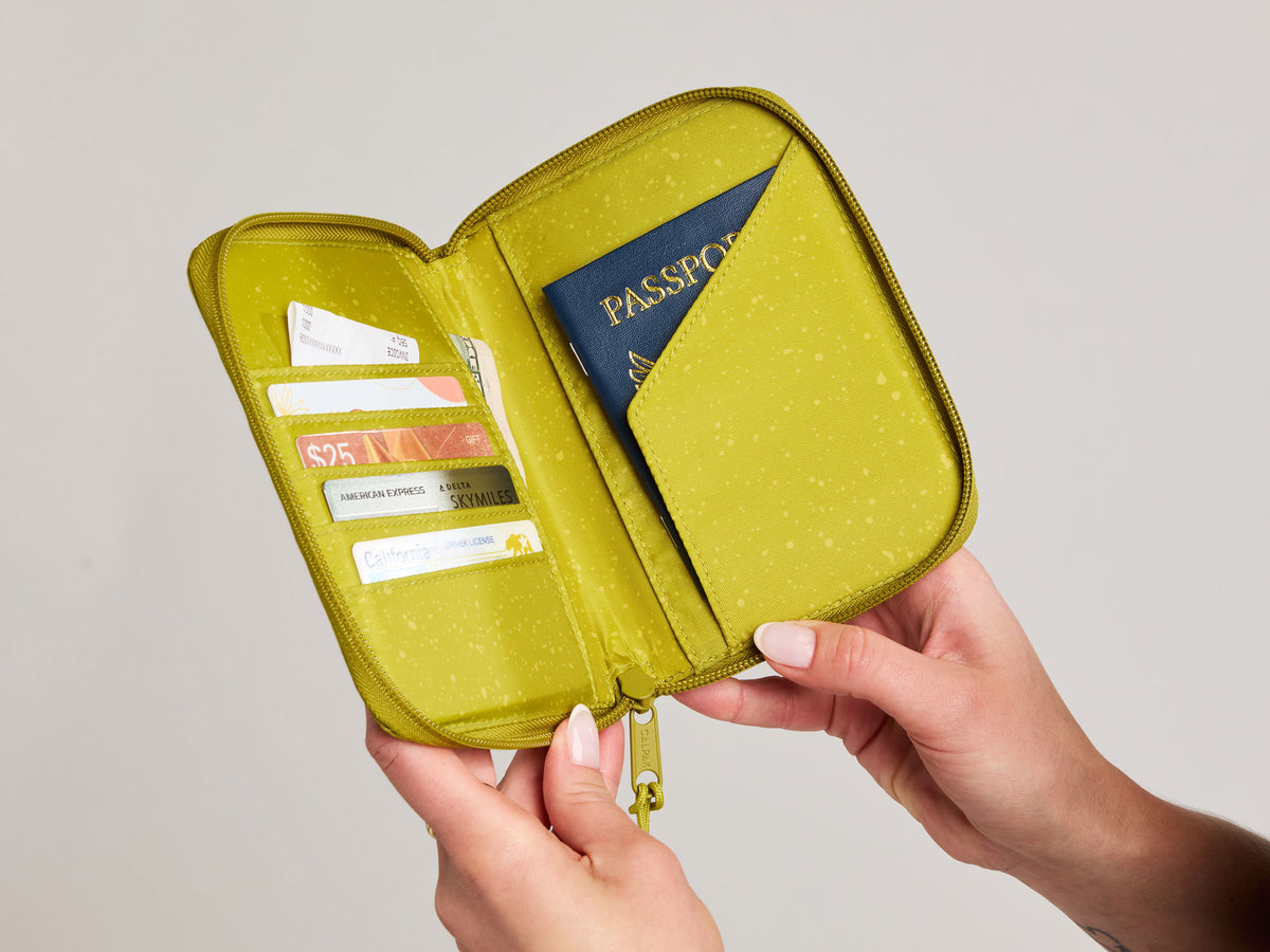 A lime green Terra Zippered Passport  Wallet holder case held open by two hands, showing its interior compartments filled with cards and a passport, against a neutral light gray background.