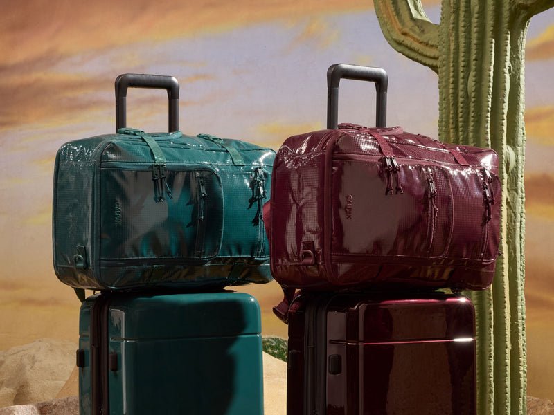 Two glossy duffel bags stacked on top of hard-shell suitcases, set against a desert backdrop with a cactus. One bag is teal and the other is burgundy, with matching suitcase colors below.