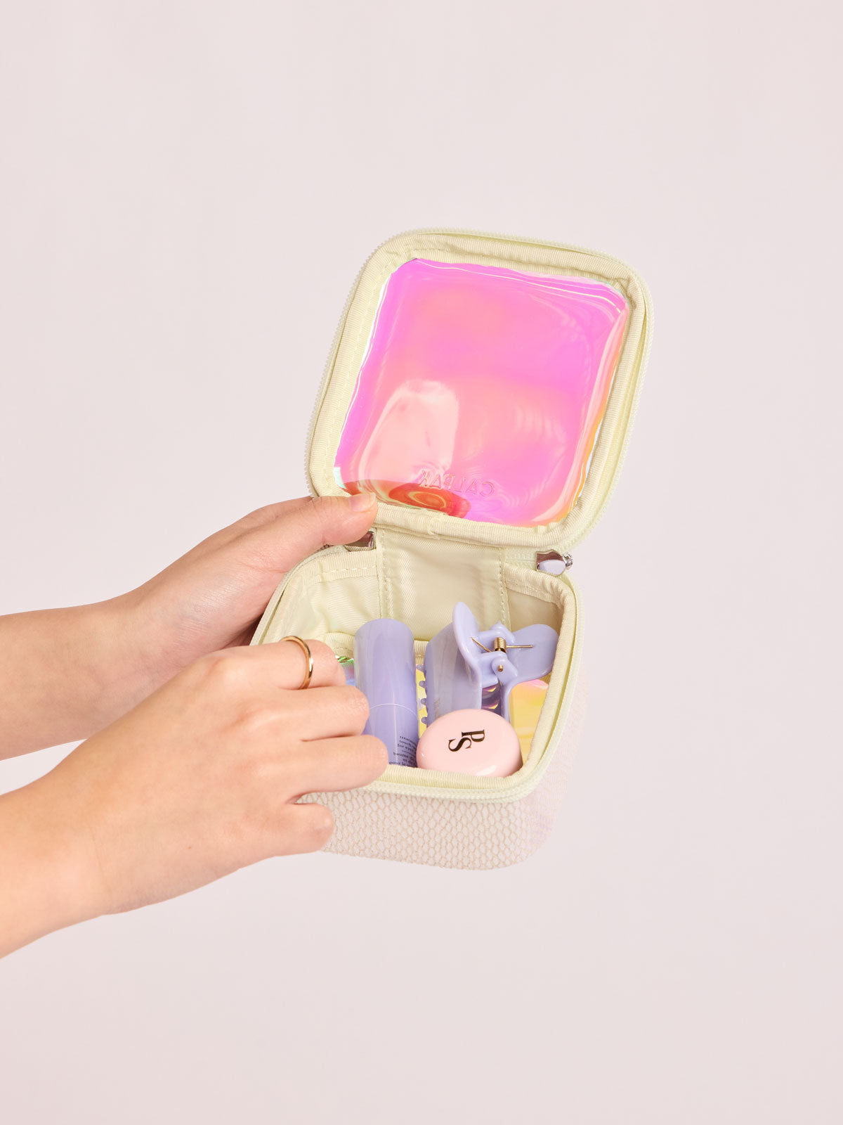 Model adding items into the CALPAK Tiny Clear Cometic Case that is square-shaped in iridescent with pink in Moonbeam with a white border on a light gray background; CCT2501-MOONBEAM