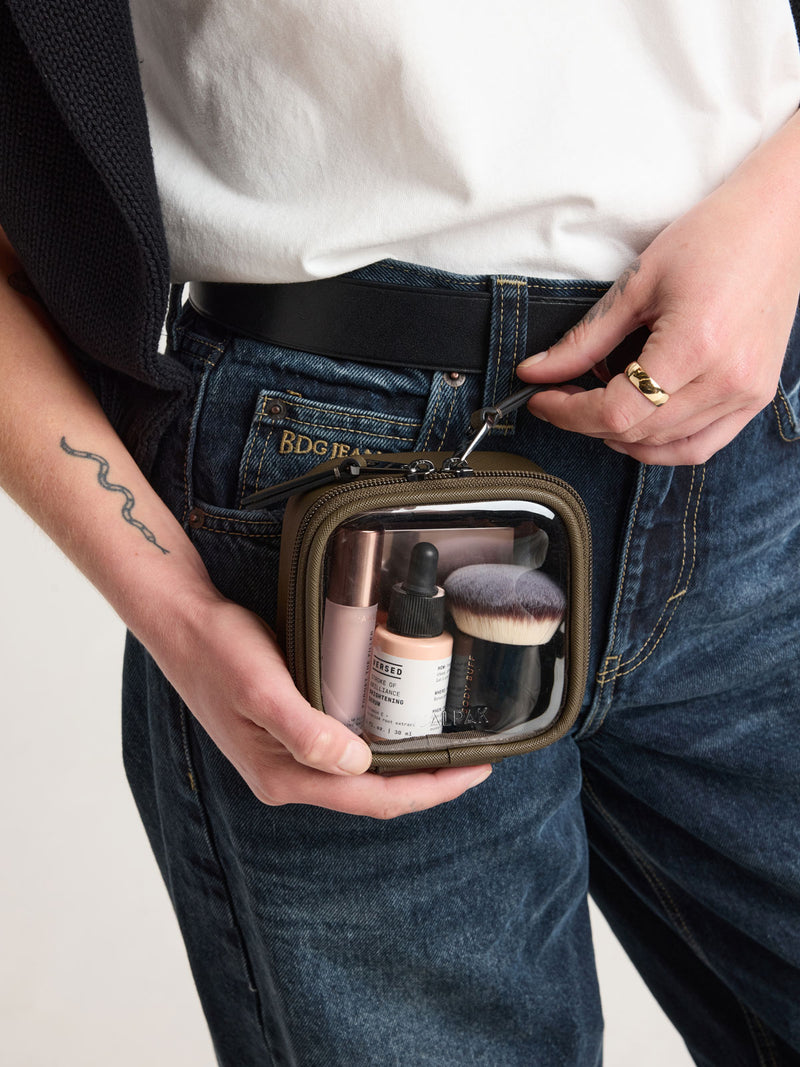 Person zipping open CALPAK Tiny Clear Cosmetic Case in brown Mushroom