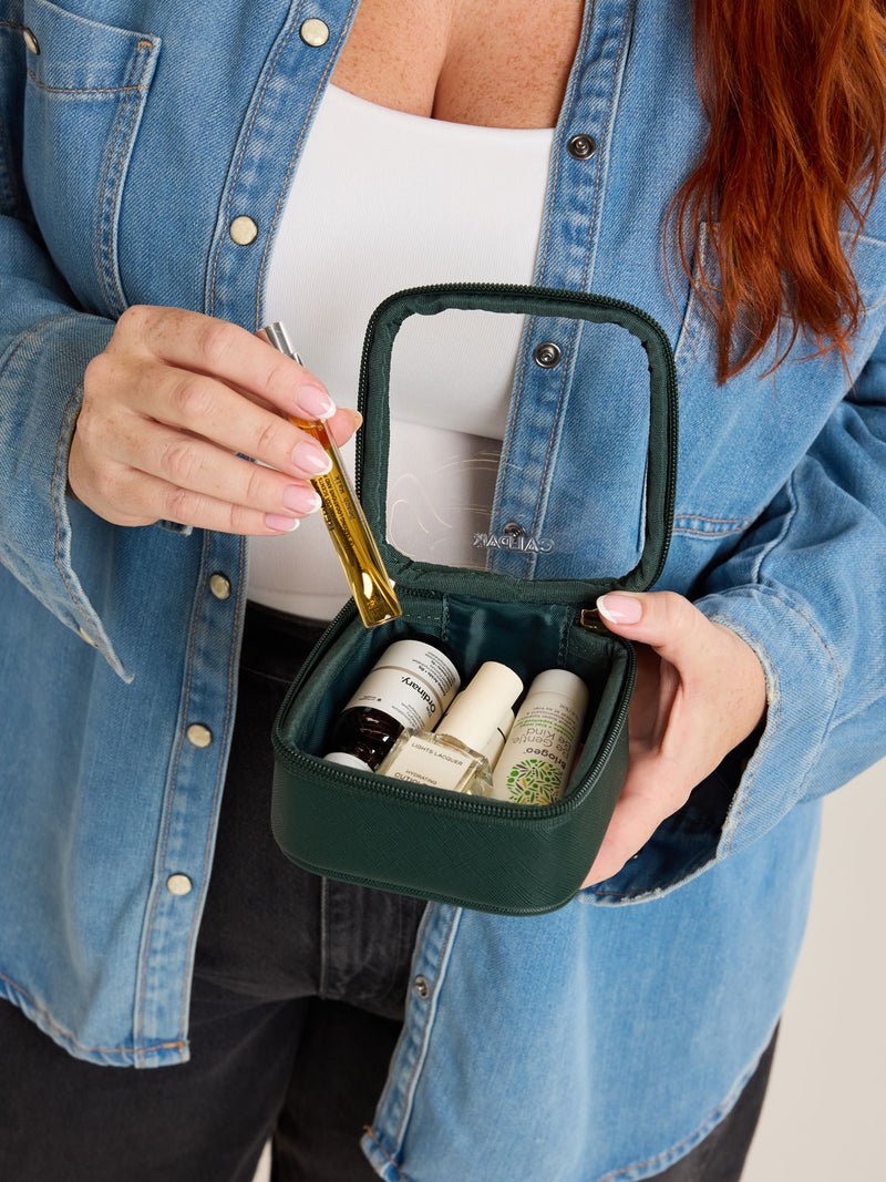 Model putting in items into CALPAK Tiny Clear Cosmetics Case in dark green Emerald on a white background