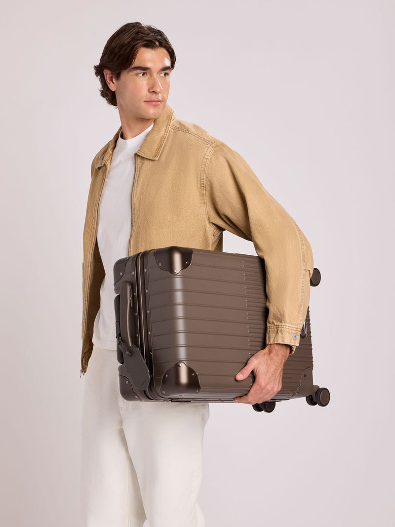 A man in a beige jacket and white shirt holds a CALPAK Vault brown Mushroom hard-shell spinner Carry On Luggage with a telescoping handle extended, featuring a black leatherette side handle and trim, set against a plain light gray background