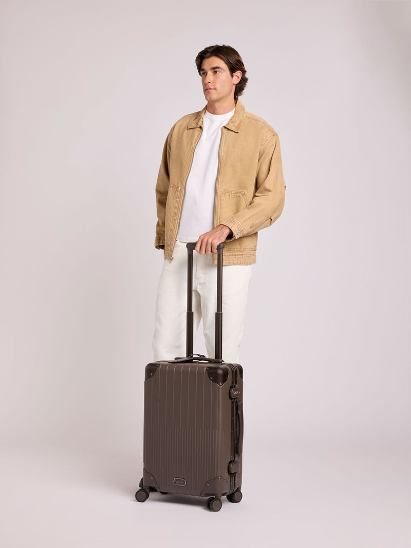 A man wearing a beige jacket, white t-shirt, and white pants stands next to a CALPAK Vault brown Mushroom hard-shell spinner Carry On Luggage with a telescoping handle extended, featuring a black leatherette side handle and trim, set against a plain light gray background