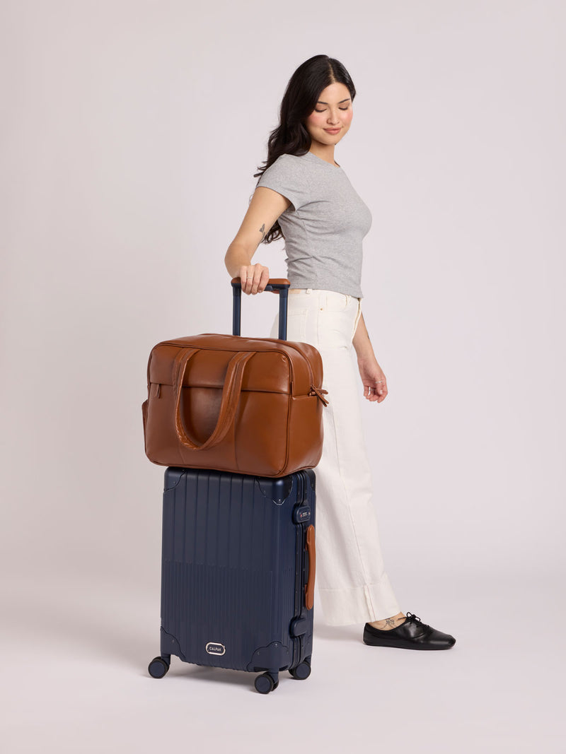 A woman stands holding the handle of a CALPAK Vault Navy blue hard-shell spinner Carry On Luggage, with a brown vegan leather Luka Duffel bag stacked on top. She is wearing a gray t-shirt, white pants, and black shoes against a light gray background