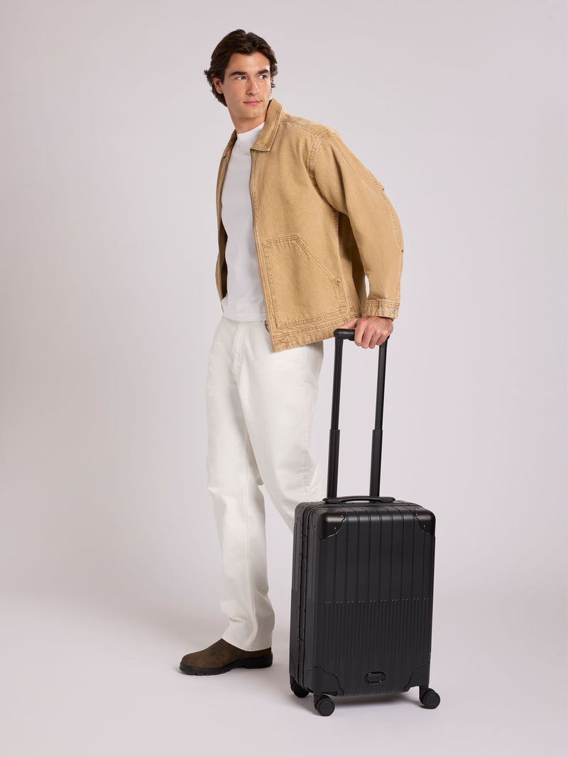 A man stands wearing a tan overshirt, white t-shirt, white trousers, and brown shoes, holding the handle of a CALPAK Vault black Onyx hard-shell spinner Carry On Luggage with a telescoping handle extended, featuring a Black leatherette side handle and trim, set against a plain light gray background