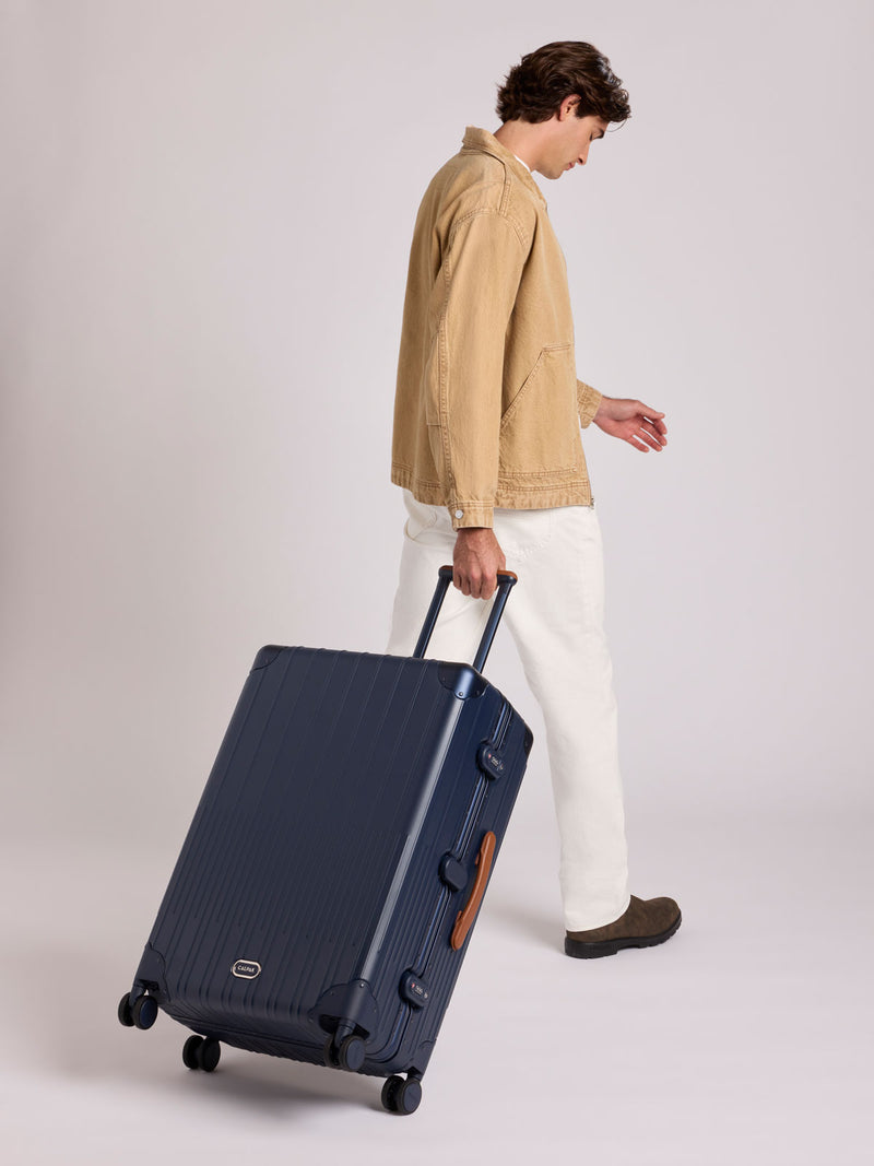 A man in a beige jacket and white pants walks while pulling a CALPAK Large Vault Luggage in blue Navy with hard-shell spinner suitcase with a ribbed vertical design, shown from the front on a light gray background