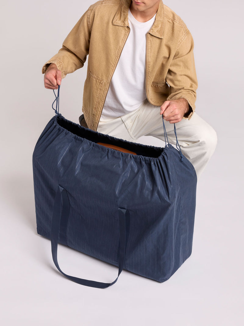 A person wearing a tan jacket, white t-shirt, and light pants holds open a large navy blue storage bag with drawstring handles of CALPAK Large Vault Luggage in blue Navy with hard-shell spinner suitcase with a ribbed vertical design, shown from the front on a light gray background