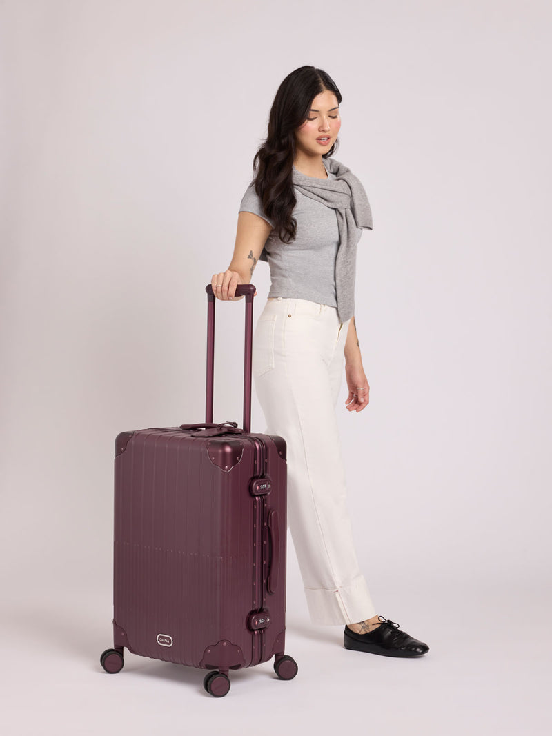 A woman wearing a grey top stands beside a CALPAK Vault Medium Luggage Dark Cherry deep red hard-shell spinner suitcase with a ribbed vertical design, shown from the front against a clean white background