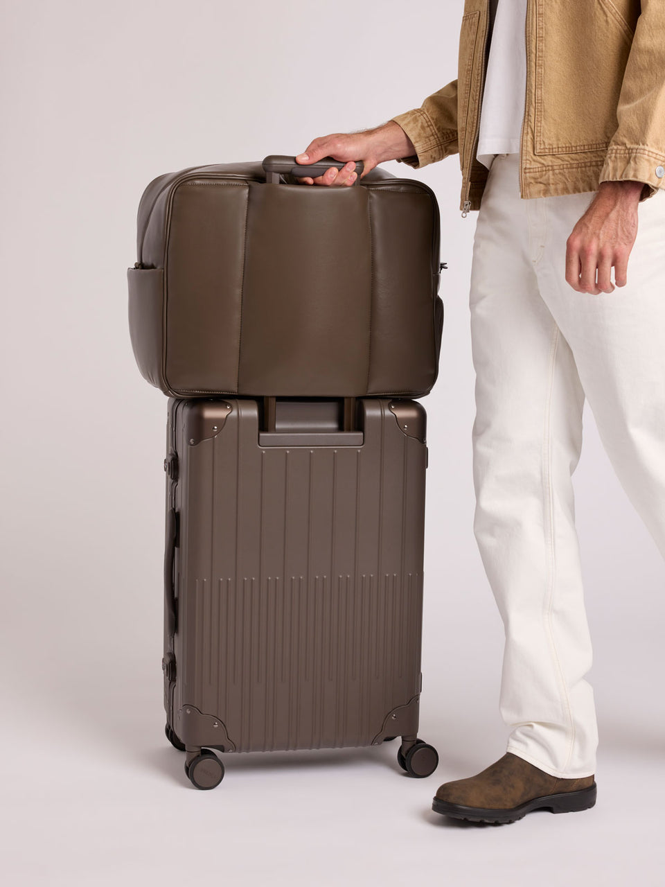 A person wearing a tan jacket, white pants, and brown shoes stands beside a CALPAK Vault luggage brown Mushroom hard-shell spinner suitcase with a ribbed vertical design, shown from the front against a clean white background; LVT1024-MUSHROOM