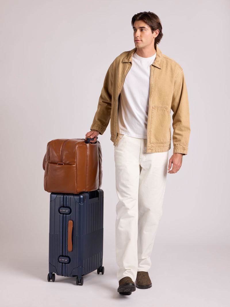 A man stands in a studio wearing a tan overshirt, white T-shirt, white trousers, and brown shoes, holding the handle of a CALPAK Vault Luggage blue Navy hard-shell spinner suitcase with a ribbed vertical design, shown from the front against a clean white background