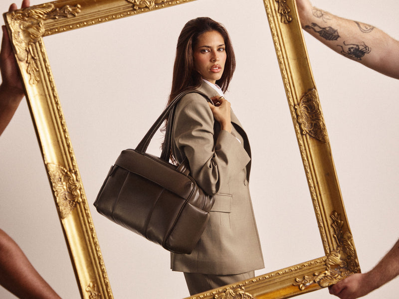 A woman in a beige blazer carries a dark brown vegan leather CALPAK Luka Mini Duffel bag, framed by a large ornate gold picture frame held by visible arms against a light beige background.